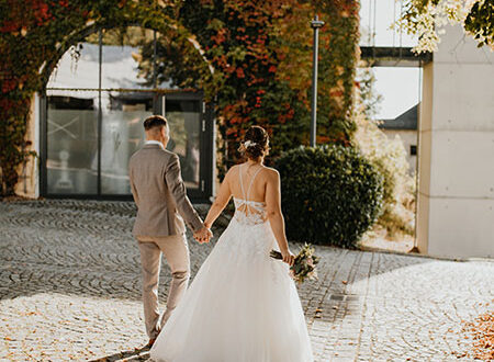 Una pareja de novios camina de espaldas por una hacienda con muros antiguos y exuberantes plantas. La novia lleva un vestido blanco con cola mediana y sostiene su ramo de flores mientras avanzan juntos hacia su boda