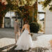 Una pareja de novios camina de espaldas por una hacienda con muros antiguos y exuberantes plantas. La novia lleva un vestido blanco con cola mediana y sostiene su ramo de flores mientras avanzan juntos hacia su boda