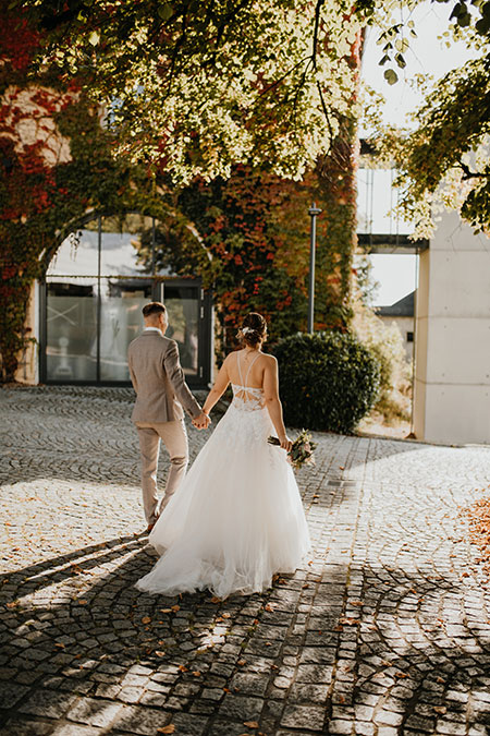 Una pareja de novios camina de espaldas por una hacienda con muros antiguos y exuberantes plantas. La novia lleva un vestido blanco con cola mediana y sostiene su ramo de flores mientras avanzan juntos hacia su boda